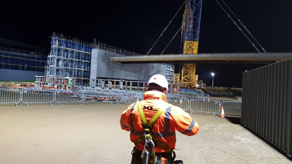 Night-time site, floodlit, with an operative in orange hi-vis watching a crane lift alongside a scaffolded building